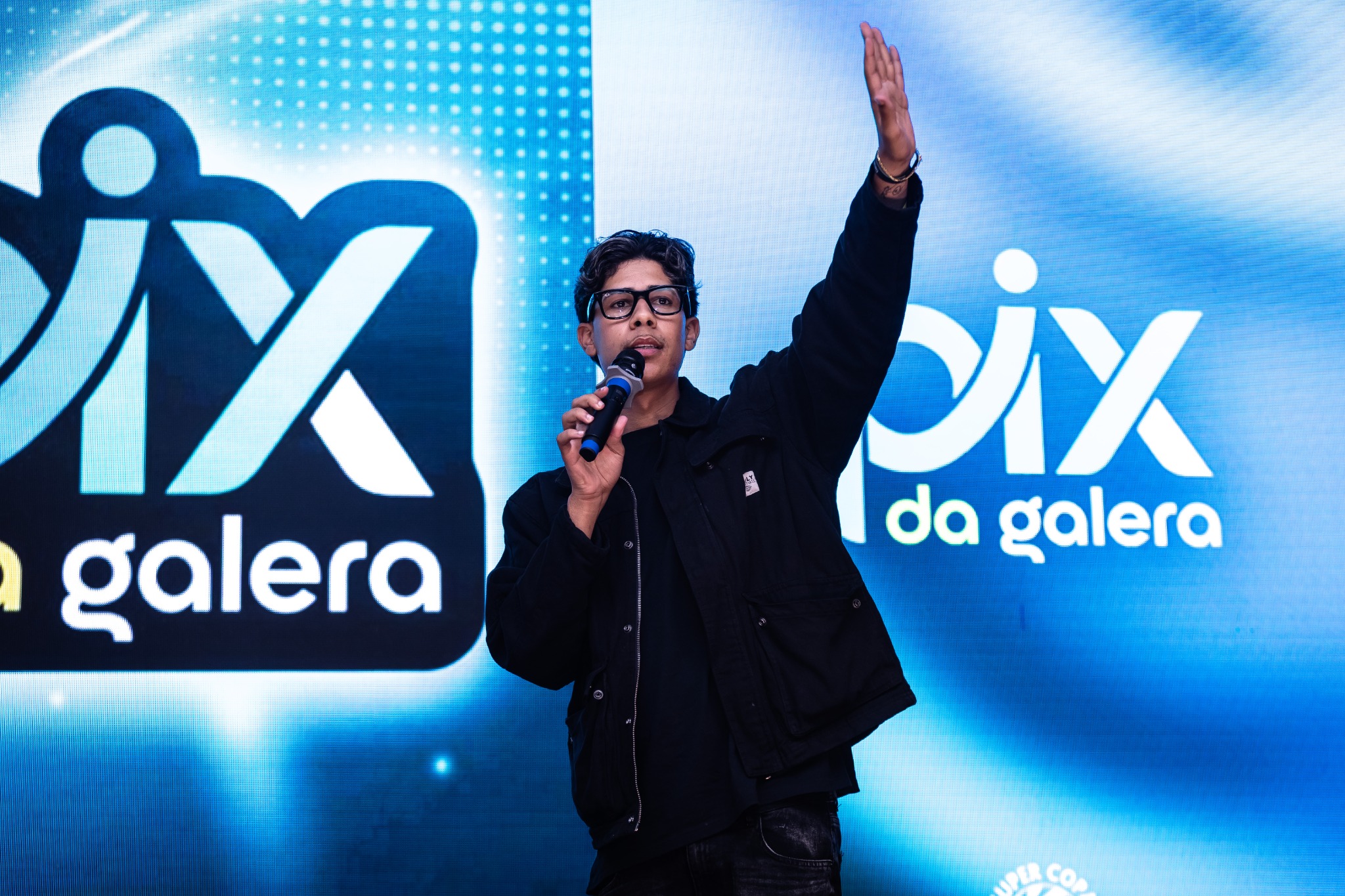 Apresentador com microfone diante do painel da PIX DA GALERA.