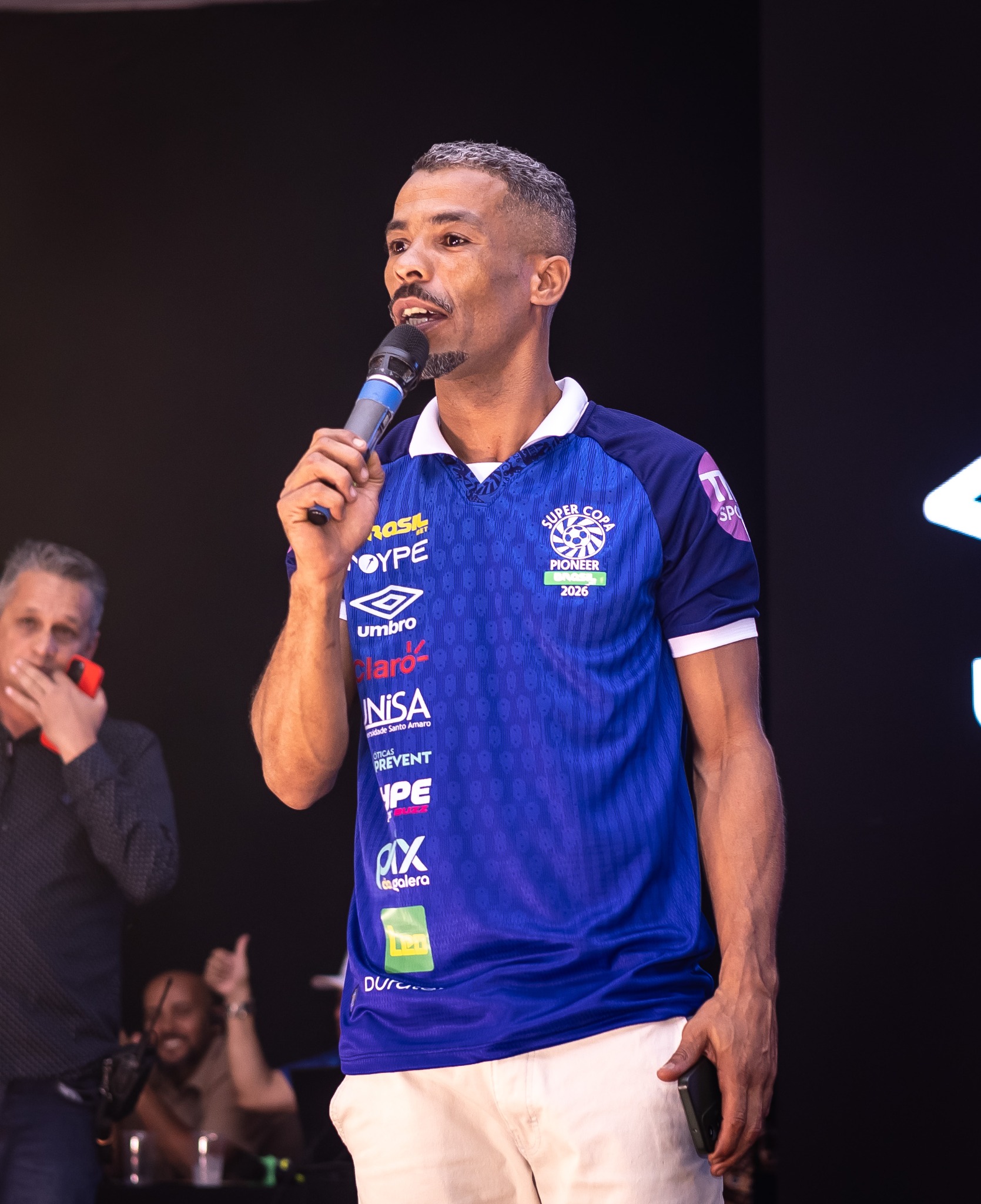 Convidado fala ao público usando uma camisa azul da competição.