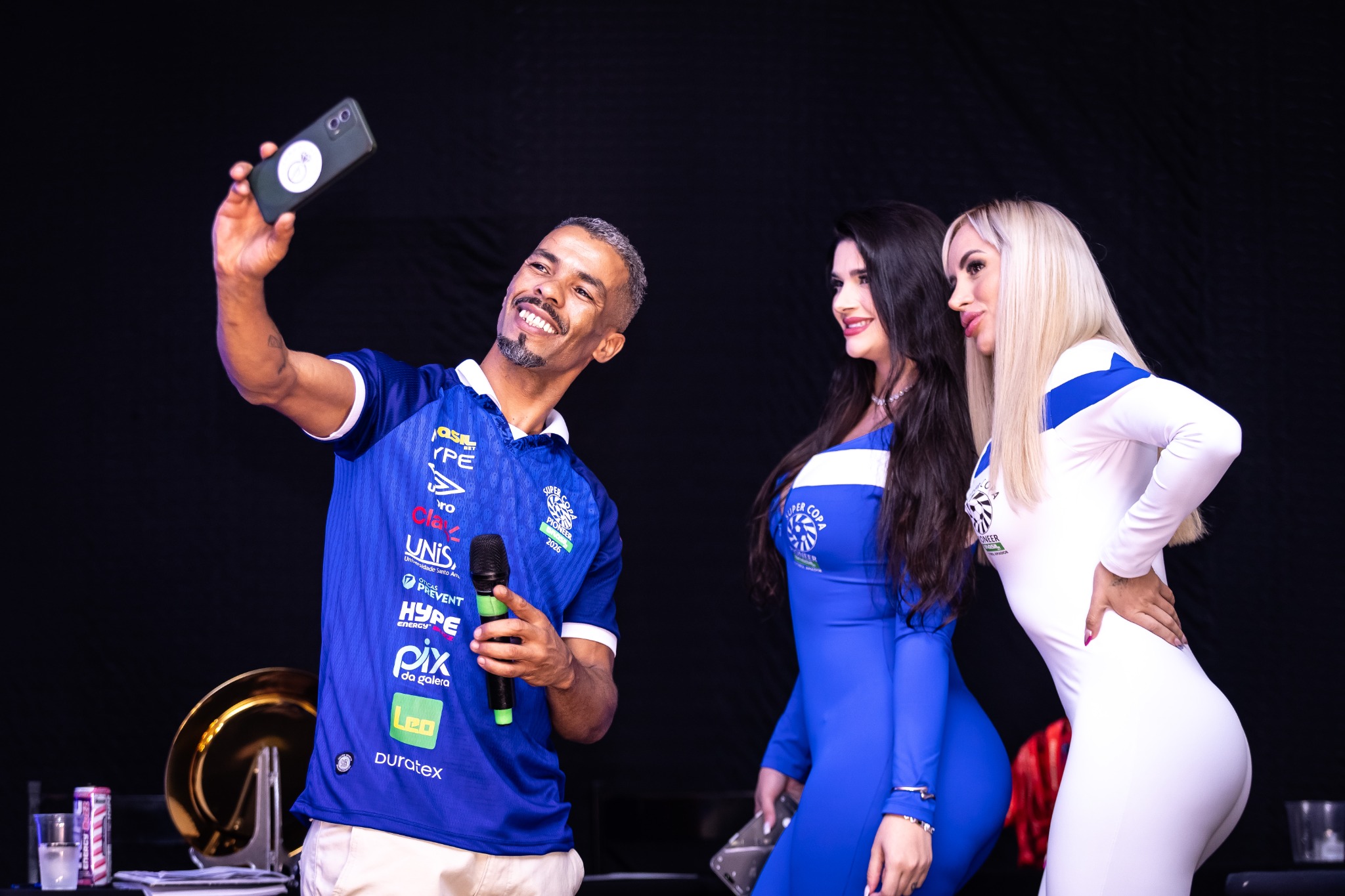 Apresentador e participantes no palco durante um momento com o celular em destaque.