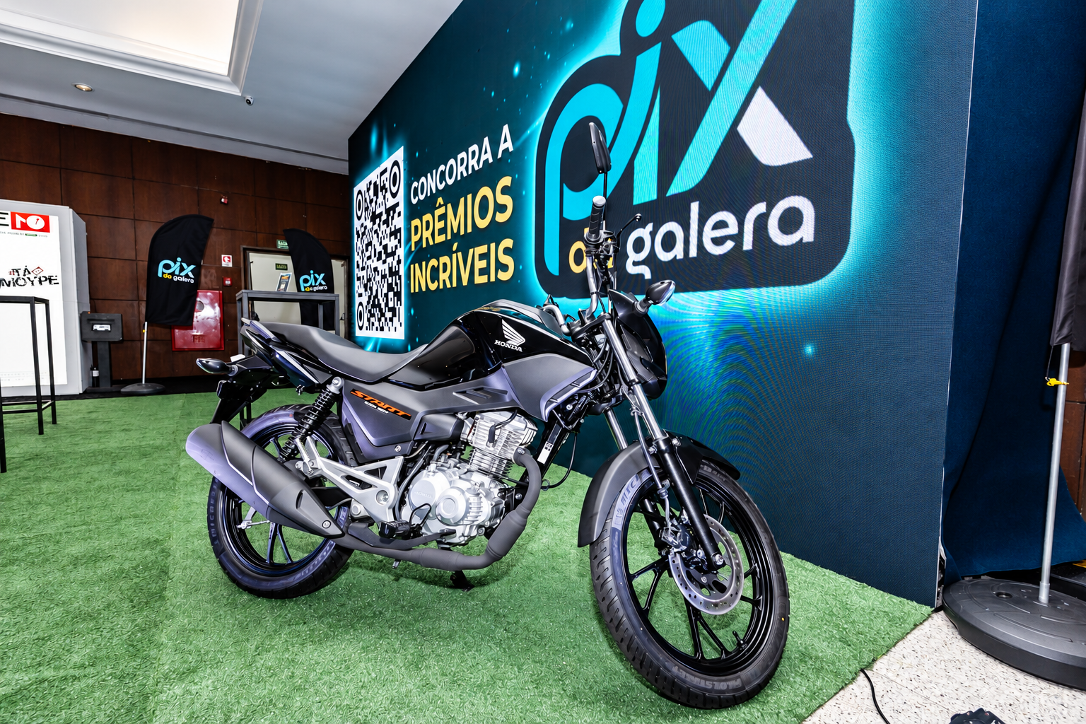 Motocicleta exposta no espaço da PIX DA GALERA, diante de painel digital azul e sinalização do evento