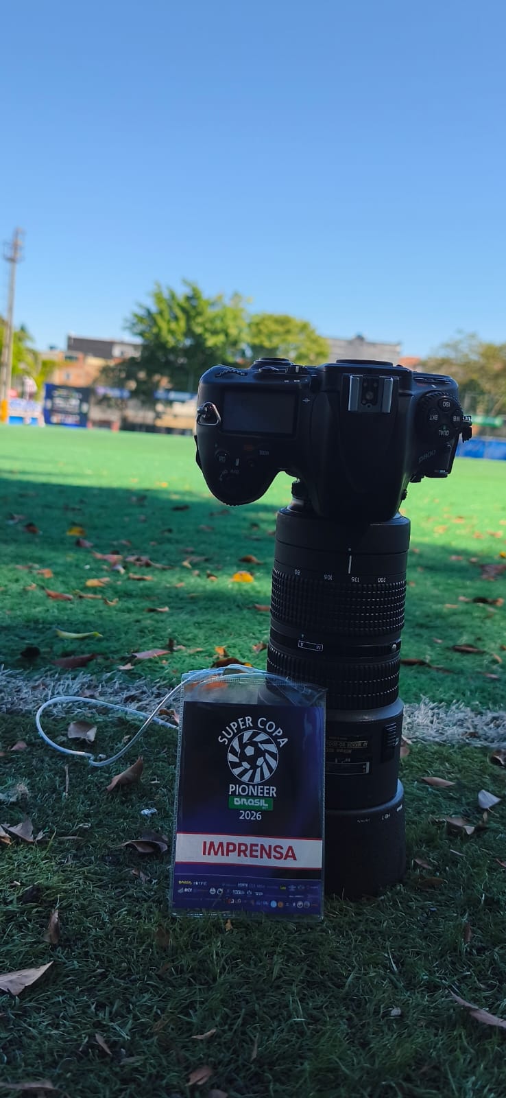Câmera fotográfica e credencial de imprensa da Super Copa Pioneer 2026 junto ao campo