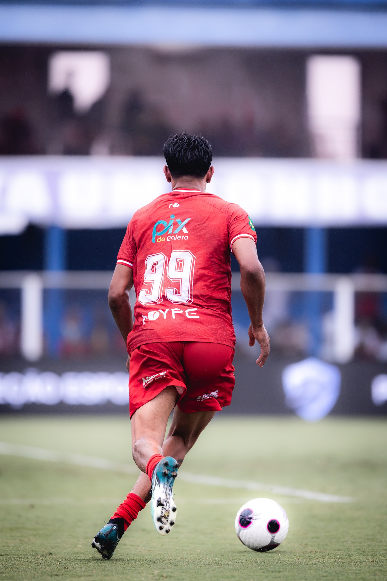 Jogador conduz a bola durante a partida; a marca aparece nas costas do uniforme vermelho.