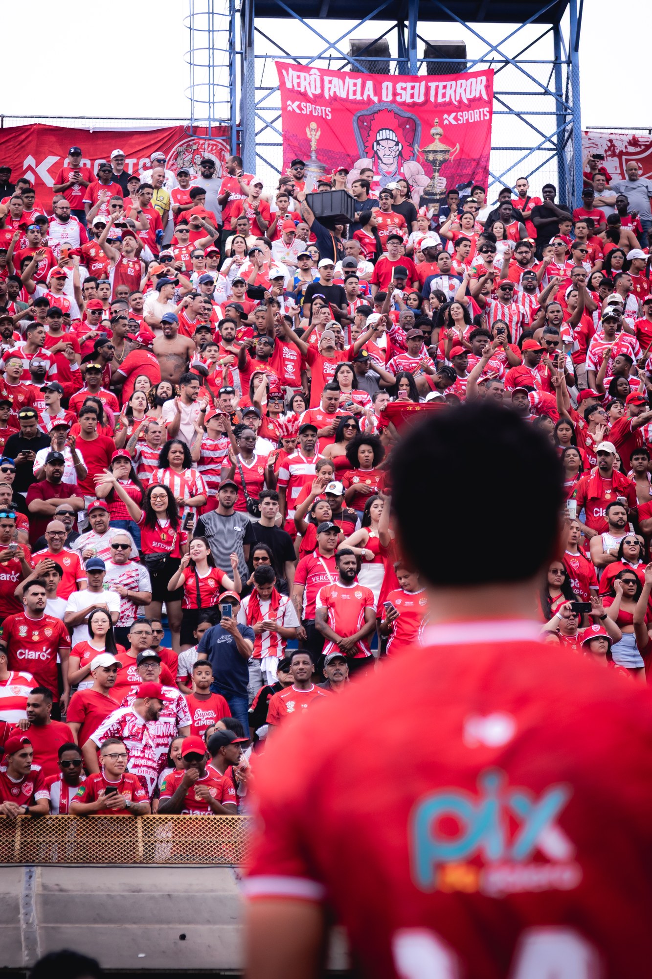 Torcida acompanha a partida; jogador em primeiro plano mostra a marca no uniforme.